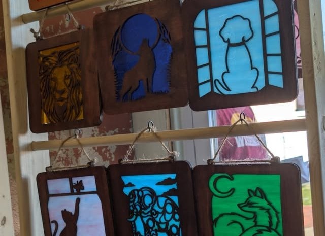 Laser-cut wood and glass suncatchers hanging on a display: lion, howling wolf, dog in a window, cat, fox